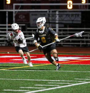 Lacrosse players in action under stadium lights; one in black uniform dodges an opponent in white during a game.