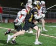 Lacrosse players compete on a field, with one athlete dodging an opponent while holding a lacrosse stick.