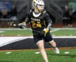 Lacrosse player in black and yellow uniform running on the field, holding a stick with focused determination.