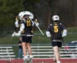 Two lacrosse players celebrating on the field, one jumping in excitement while the other runs beside.