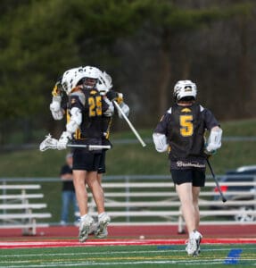 Two lacrosse players celebrating on the field, one jumping in excitement while the other runs beside.