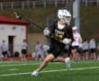 Lacrosse player in black jersey runs on field, stick in hand, during an evening practice session.