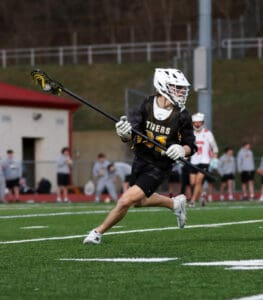 Lacrosse player in black jersey runs on field, stick in hand, during an evening practice session.