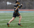 Player in black lacrosse uniform running on field, carrying a lacrosse stick, ready for action in a dynamic game.