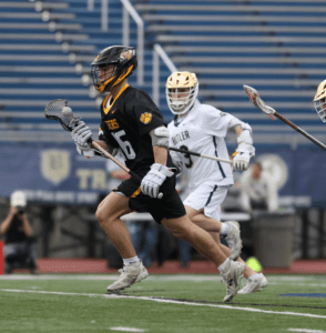 High school lacrosse players sprint on the field during a competitive match in athletic gear.