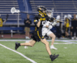 Boy in a black lacrosse uniform runs with a stick, competing in a game on a field. The action captures youth sports excitement.