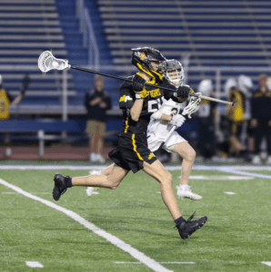 Boy in a black lacrosse uniform runs with a stick, competing in a game on a field. The action captures youth sports excitement.