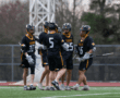 Lacrosse team in black and yellow jerseys celebrates on the field, showcasing teamwork and sportsmanship during a game.