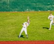 A baseball player wearing a white uniform leaps to catch a fly ball, with a teammate in the background.