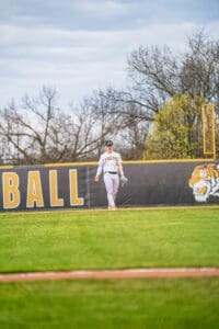 Baseball player in striped uniform walks on field with “Tigers” and “Baseball” signage in background.