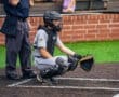 Baseball catcher in gray gear crouches behind the plate, while an umpire observes the game in the background.