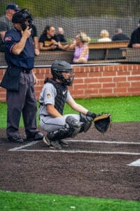 Baseball catcher in gray gear crouches behind the plate, while an umpire observes the game in the background.