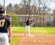 A young baseball player throws the ball to first base during a game on a sunny day. Players and a field are visible.