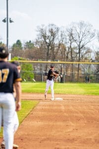 A young baseball player throws the ball to first base during a game on a sunny day. Players and a field are visible.