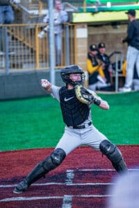 A young baseball catcher in gear prepares to throw a ball during a game on a green and red field.