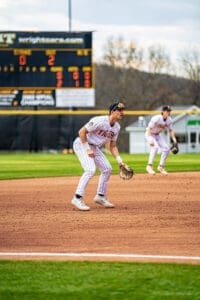 Two baseball players in striped uniforms prepare for action on a diamond, with a scoreboard showing game stats in the background.