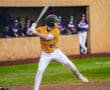 Baseball player in yellow jersey swings at pitch on a field, with teammates watching from the dugout in the background.