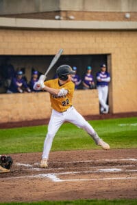 Baseball player in yellow jersey swings at pitch on a field, with teammates watching from the dugout in the background.
