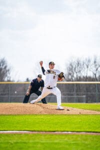 Young baseball pitcher in striped uniform throws a pitch on the mound, with the umpire in the background.