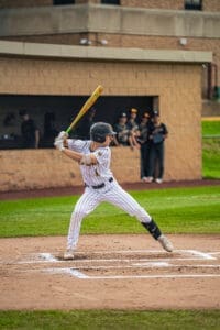 Baseball player in a striped uniform prepares to swing at the pitch, with teammates visible in the background.