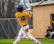A baseball player in a yellow jersey swings for a hit during a game, dirt on his pants, focused on the ball.