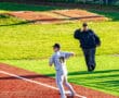 Baseball player in grey uniform throws a ball while coach watches on a vibrant green and red field during practice.