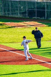 Baseball player in grey uniform throws a ball while coach watches on a vibrant green and red field during practice.