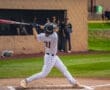 Baseball player in a pinstripe uniform swinging a bat, with spectators watching from the dugout in the background.