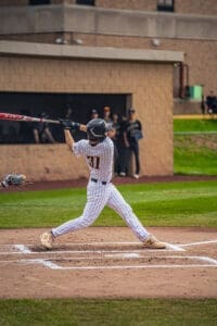 Baseball player in a pinstripe uniform swinging a bat, with spectators watching from the dugout in the background.