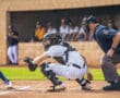 Catcher preparing to receive a pitch, with an umpire and players in the background during a baseball game.