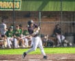 Baseball player swings bat at home plate during a game, with teammates in the dugout visible in the background.