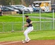 A baseball player in a black jersey prepares to pitch on a sunny field, surrounded by spectators and parked cars.