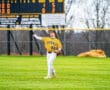 A baseball player in a yellow jersey throws a ball on the field with a scoreboard in the background.