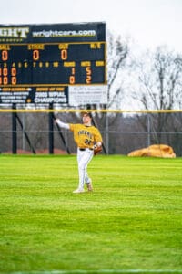 A baseball player in a yellow jersey throws a ball on the field with a scoreboard in the background.