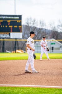 Baseball players on the field during a game, with a scoreboard in the background and a sunny day above them.