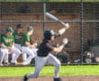Young baseball player swings at a pitch during a game, with teammates visible in the dugout behind him.