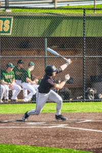 Young baseball player swings at a pitch during a game, with teammates visible in the dugout behind him.