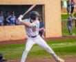 High school baseball player in pinstriped uniform prepares to bat during a game, with spectators in the background.