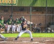 Young baseball player prepares to swing at a pitch, with teammates in the dugout during a game.
