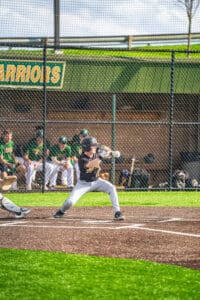 Young baseball player prepares to swing at a pitch, with teammates in the dugout during a game.