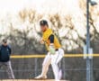 A young pitcher in a yellow jersey prepares to throw a baseball on the mound during a game, with an umpire in the background.