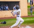 Baseball player in striped uniform swings a bat at home plate, with teammates in the background watching.