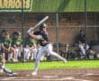 High school baseball player swings bat at home plate, with teammates in the background on a sunny day.