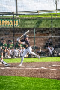 High school baseball player swings bat at home plate, with teammates in the background on a sunny day.