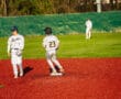 A youth baseball player in gray runs toward first base on a bright red field, while another in striped uniform looks on.