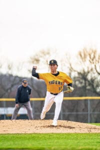 High school pitcher in a yellow Tigers uniform throws a baseball on the mound, with an umpire in the background.