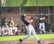 A young baseball player swings a bat during practice, with teammates visible in the background.