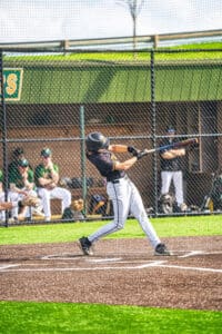 A young baseball player swings a bat during practice, with teammates visible in the background.
