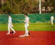 Baseball players on a bright red field, one standing on base and another walking to position in a sunny setting.