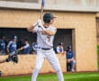 Baseball player in a white striped uniform stands ready to bat on the field with teammates in the background.